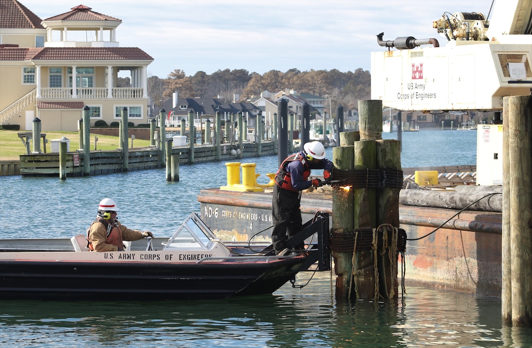 One man steers a boat while the other uses a torch to deconstruct wooden pilings in Rudee Inlet