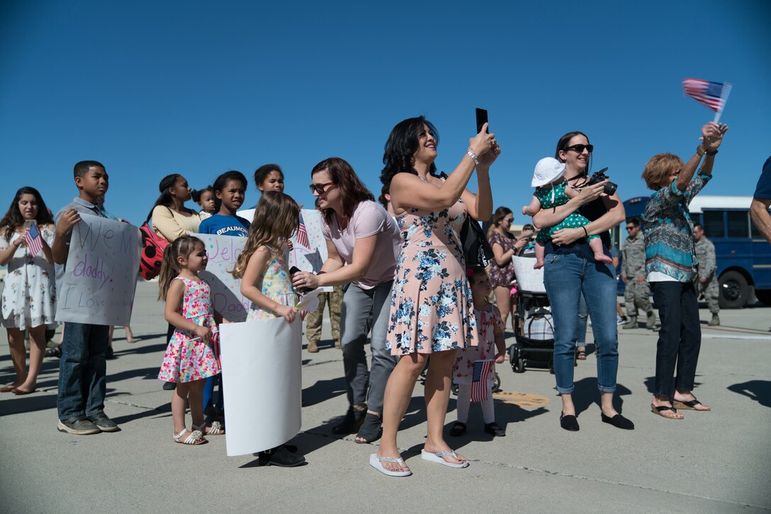 Children and women stand in a group. Many are holding cameras and carrying “Welcome Home” signs.