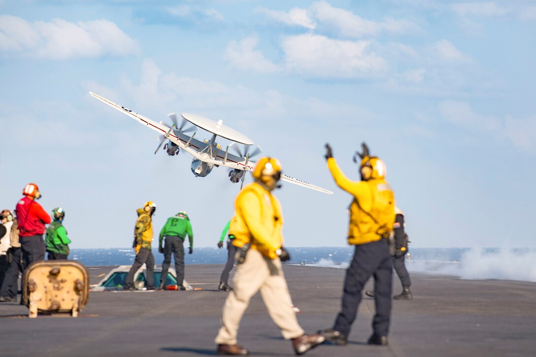 A Navy aircraft flies off of the deck of a ship as sailors watch.