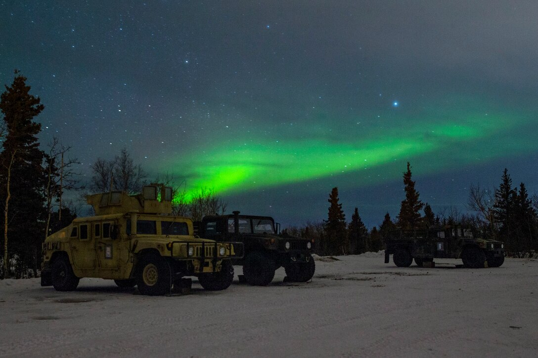 Humvees sit parked on snowy ground under a night sky with green light from an aurora