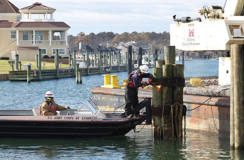 One man steers a boat while the other uses a torch to deconstruct wooden pilings in Rudee Inlet