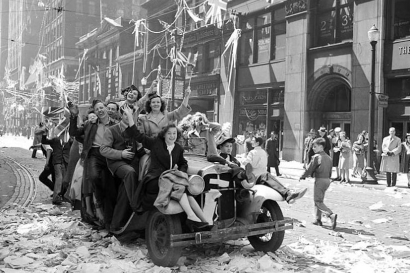 A group of people crowd into a car smiling and waving flags.