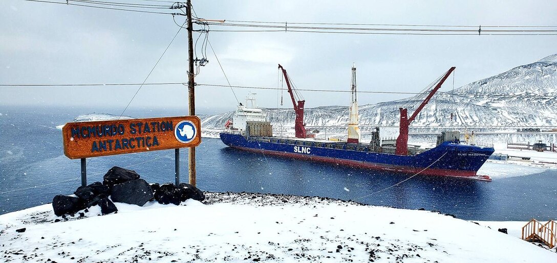 Ship docks in icy harbor. A sign in the foreground reads “McMurdo Station Antarctica.”