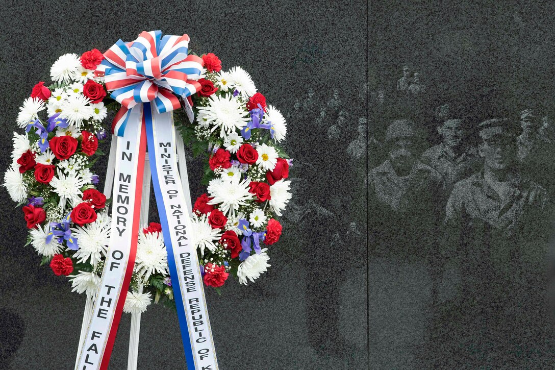 A wreath with red, white and blue flowers and memorial ribbons sits on a stand by a mural wall.