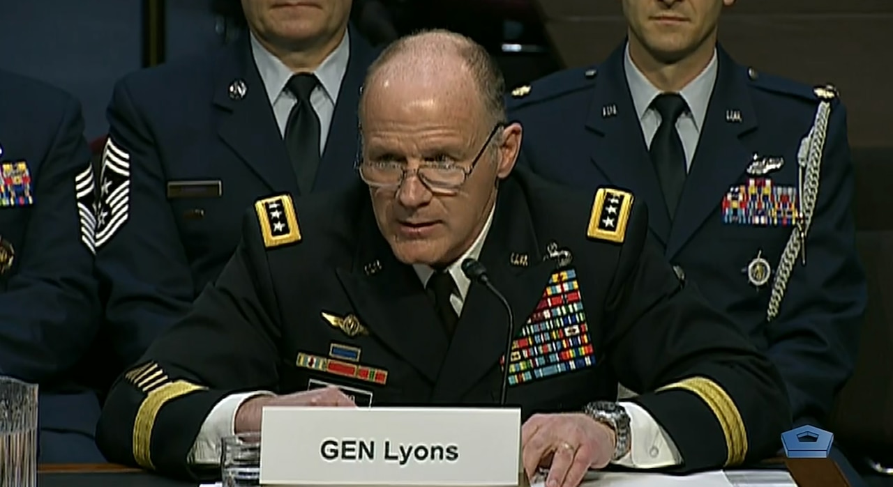 Four-star Army general seated at a table speaks into a microphone, with Air Force personnel in uniform seated behind him.