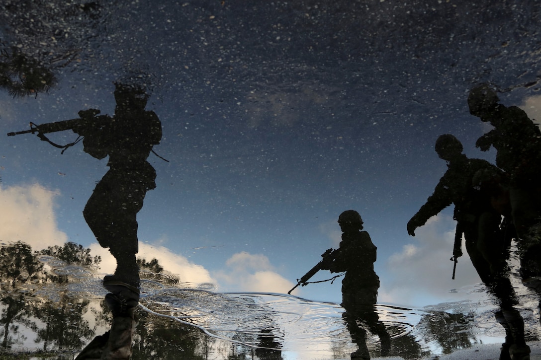 Silhouettes of Marine Corps recruits carrying weapons are reflected in water.