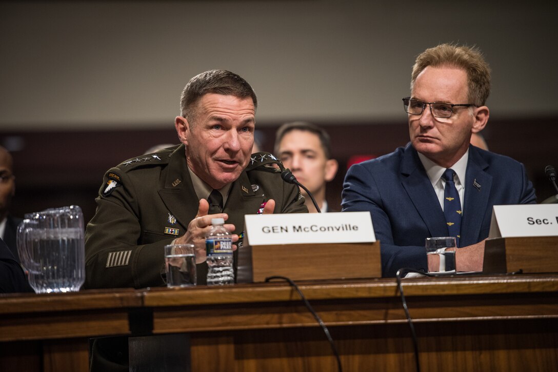A military officer speaks into a microphone while seated at a wooden table.