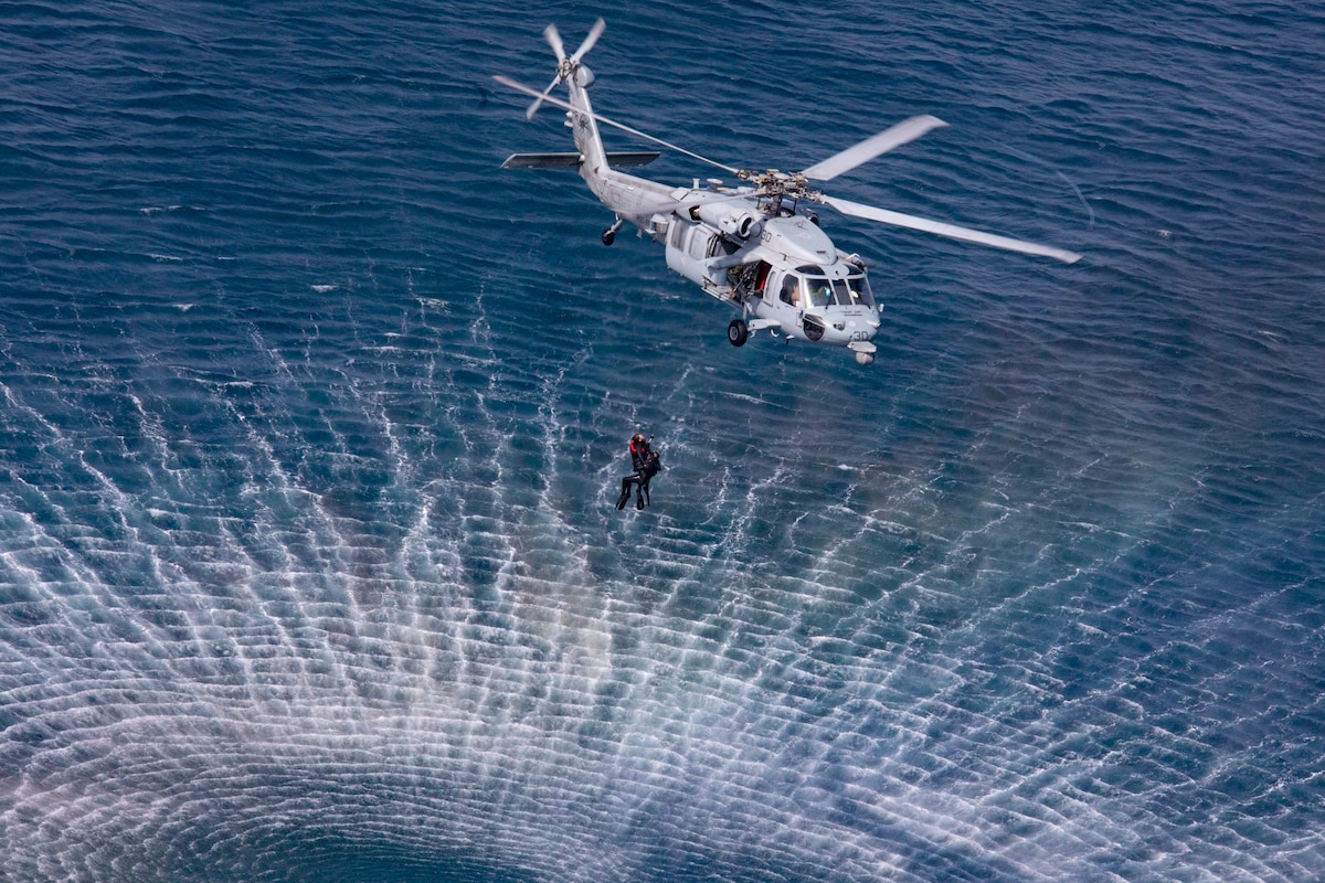 Two sailors hang on a line from a helicopter hovering over water.