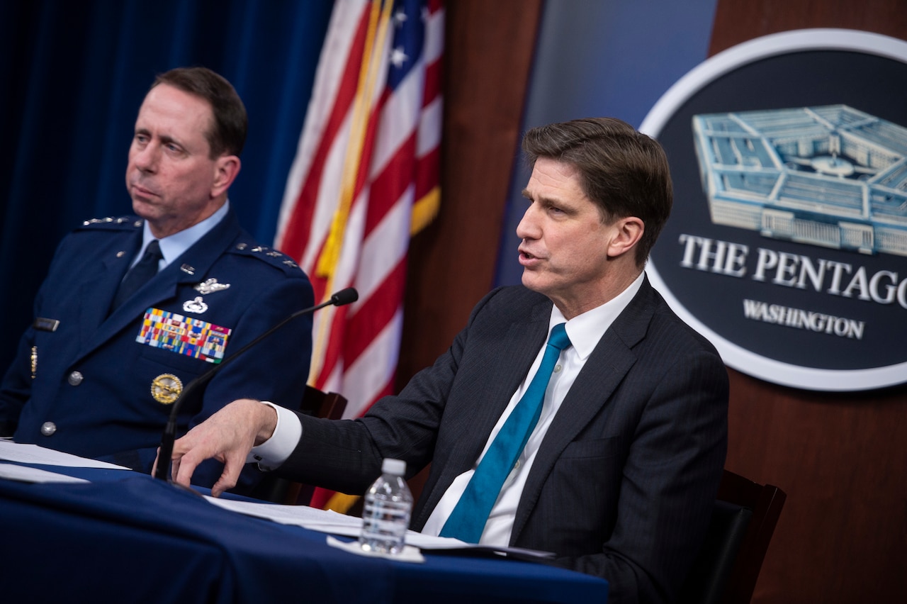 Two men sit at a table. In the background, a sign reading “The Pentagon” hangs on a wall.
