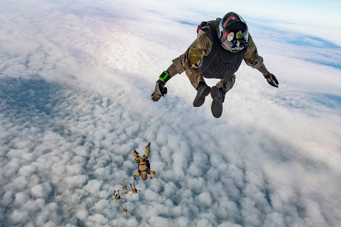 Sailors wearing parachute backpacks descend in a cloud-studded sky.