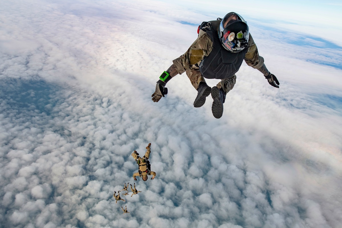 Sailors wearing parachute backpacks descend in a cloud-studded sky.