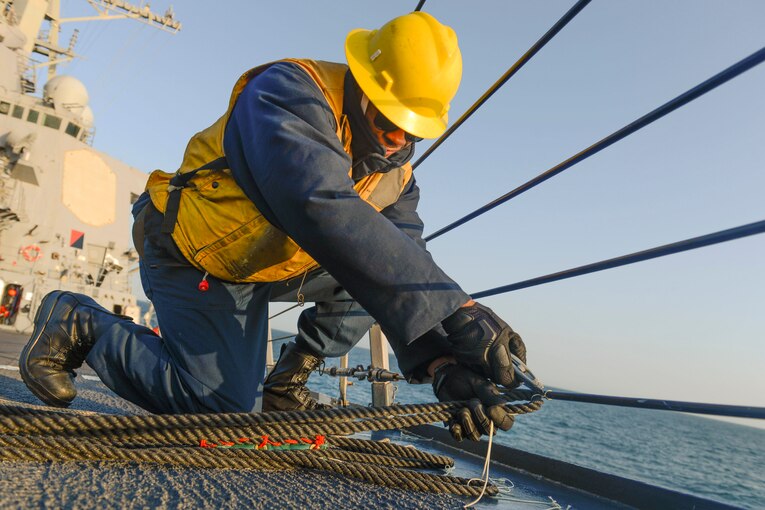 A sailor wearing a yellow hard hat and vest cuts a rope.