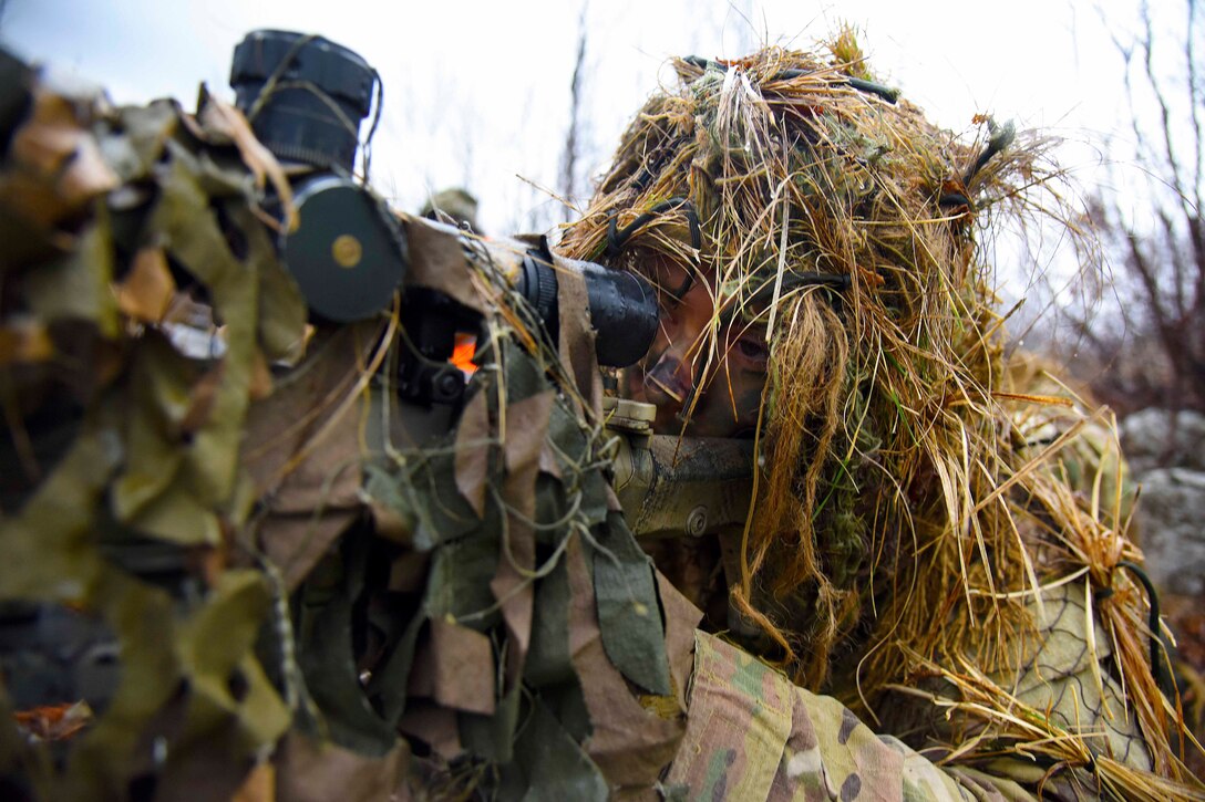 A soldier covered in grass lays on the ground aiming a weapon at a target.