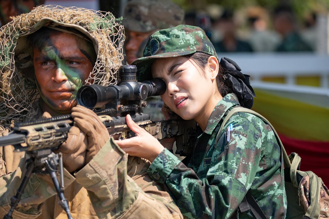A soldier aims her gun while another soldier holds it level.