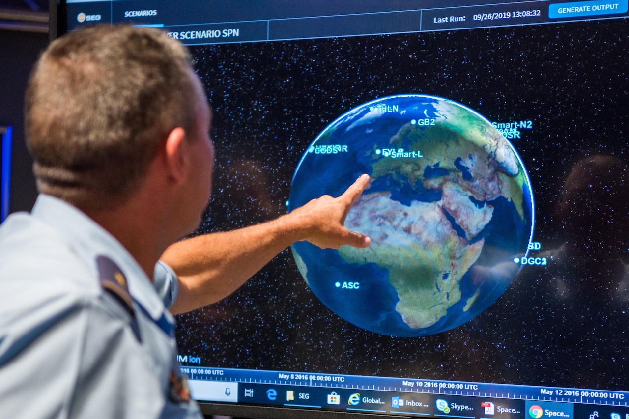 Uniformed man points at earth on wall-mounted computer screen.