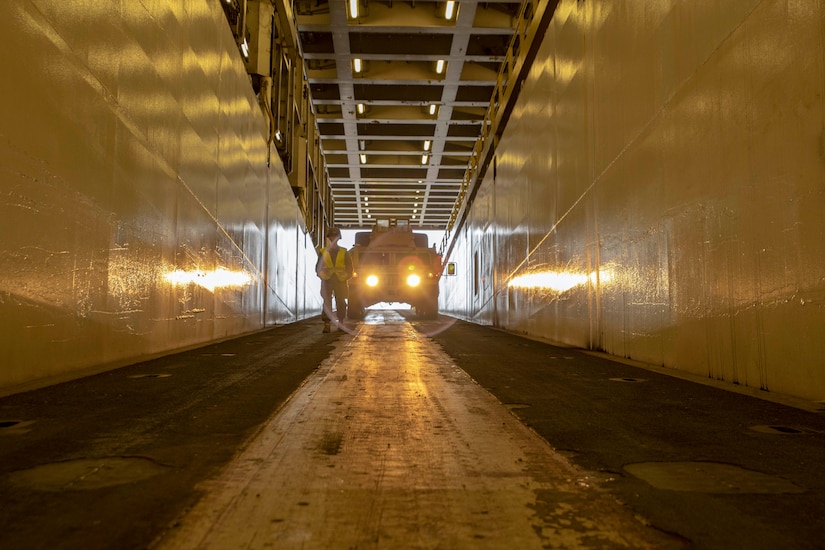 A Marine Corps vehicle is parked with headlights on aboard a cargo ship.