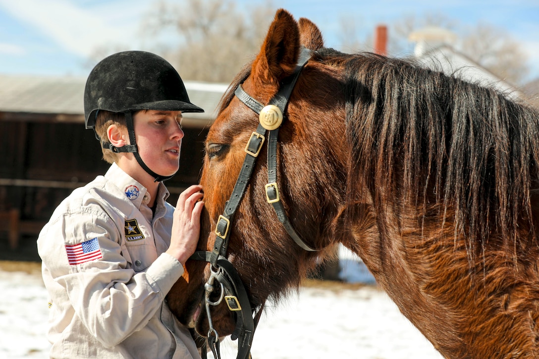 A soldier in a riding hat pets a horse's face.