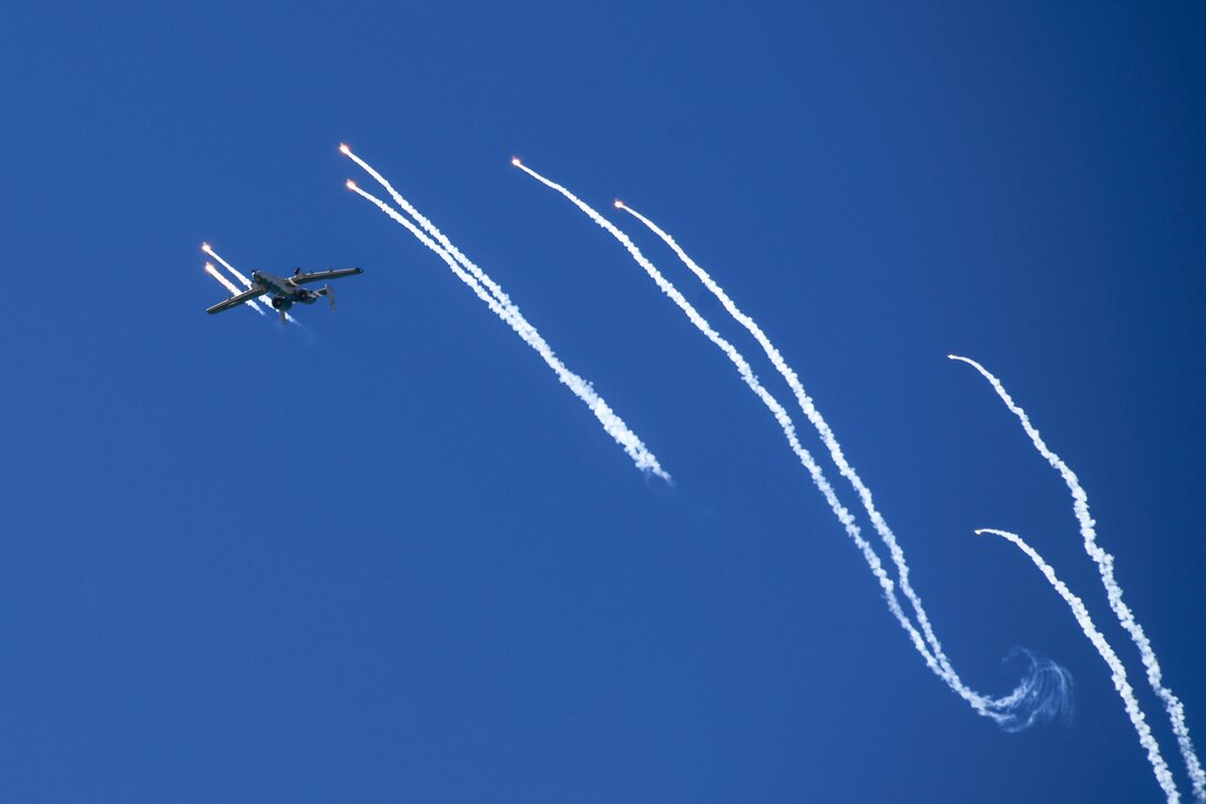 An aircraft flies in a deep blue sky streaked by white smoke stripes from flares it is releasing.