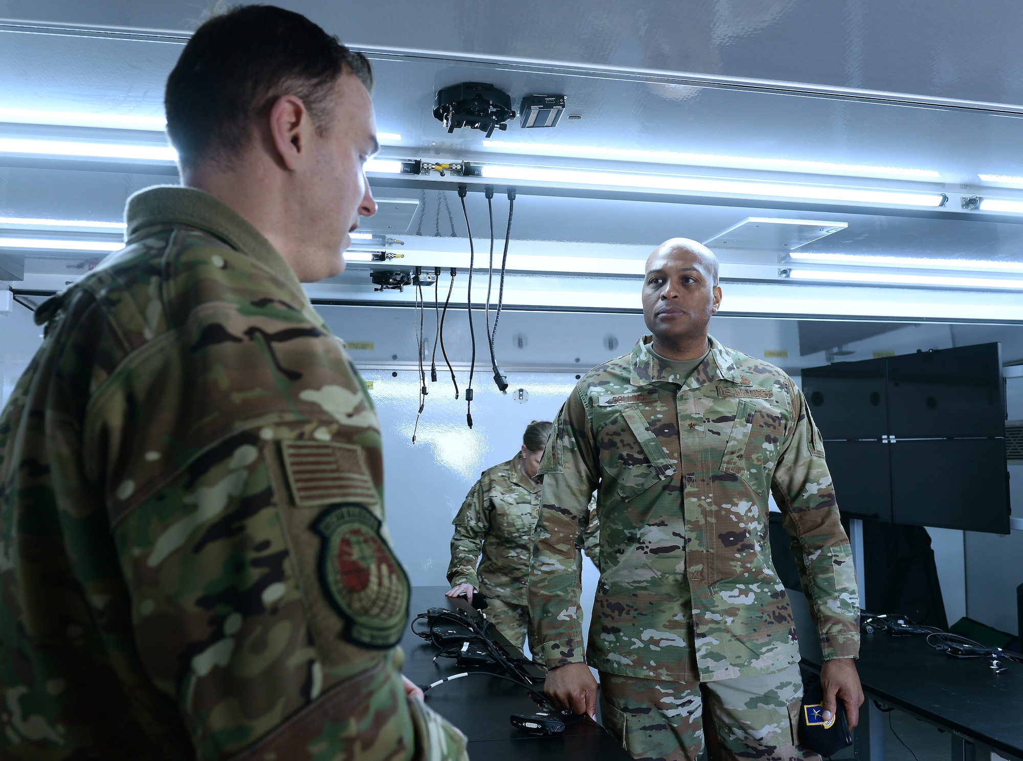 Man briefs other man in room with TV and other military member in the background