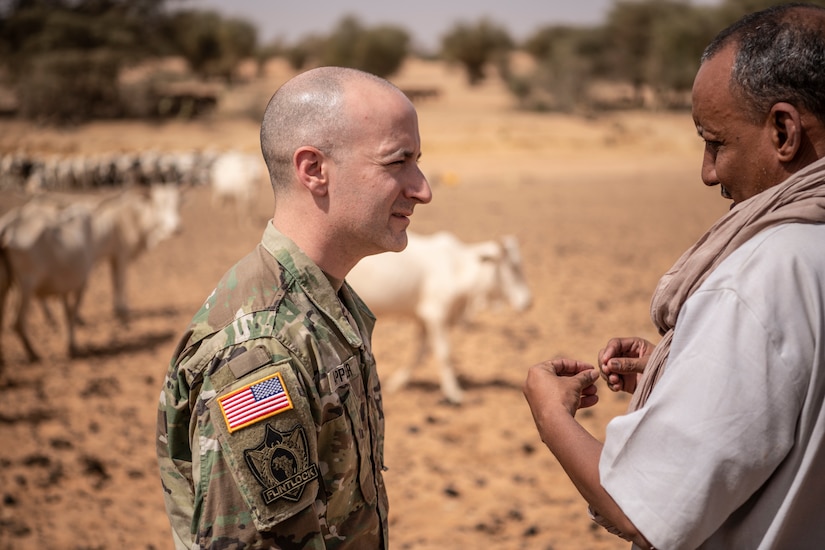 A soldier talks to a man; cattle can be seen in the background.