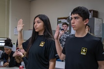 Dark-haired male and dark-haired female both in black shirts stand with right hands raised in front of people in classroom.