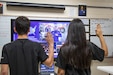 Male and female in black t-shirts with dark hair raise their right hands in front of a tv screen in a classroom.