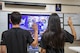 Male and female in black t-shirts with dark hair raise their right hands in front of a tv screen in a classroom.