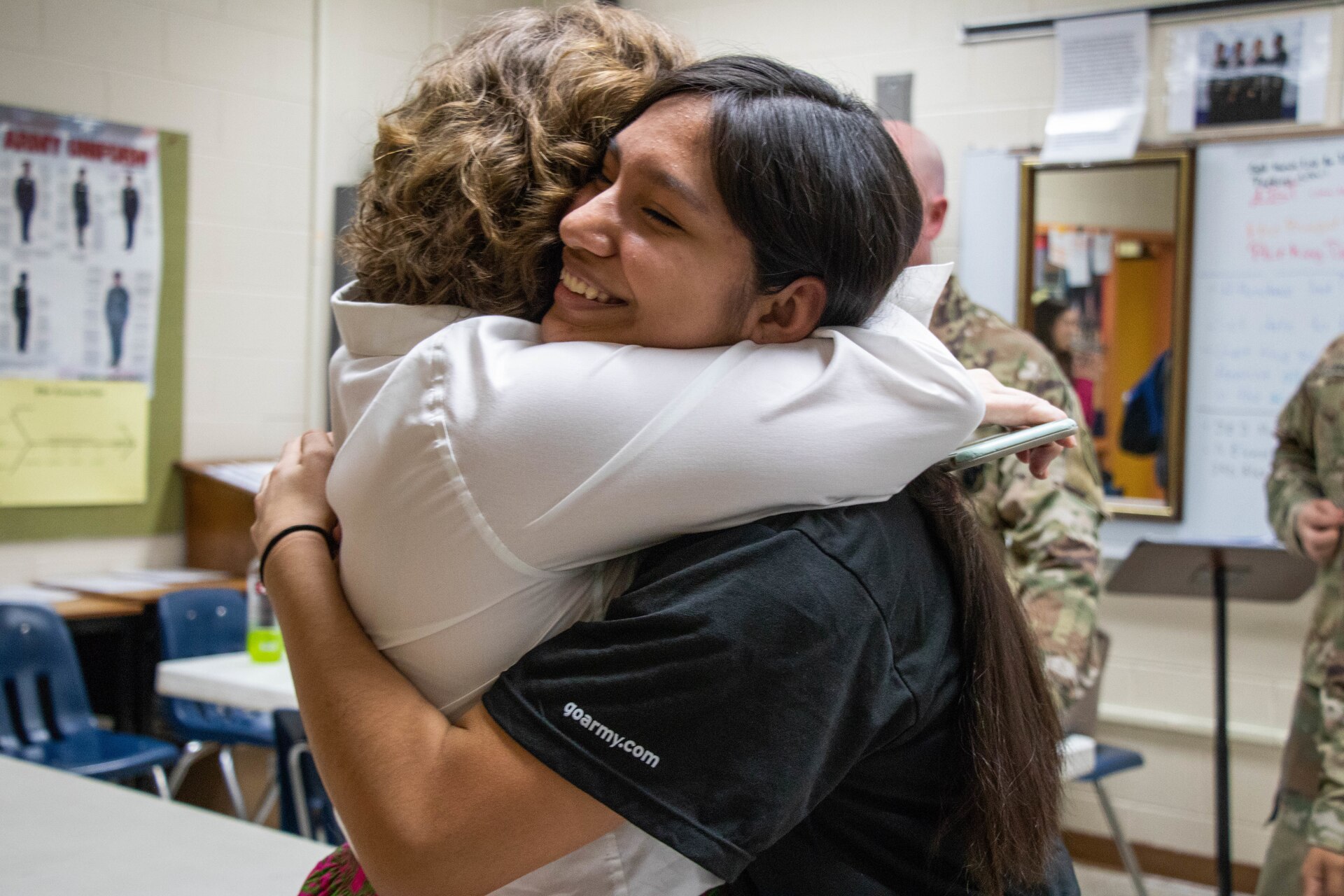 Lady in white shirt embraces young lady in black shirt in a classroom.
