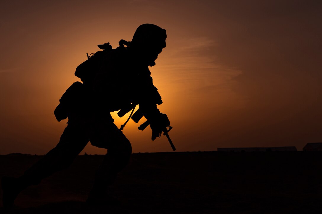 A Marine holds a weapon while the sky is lit orange and yellow.