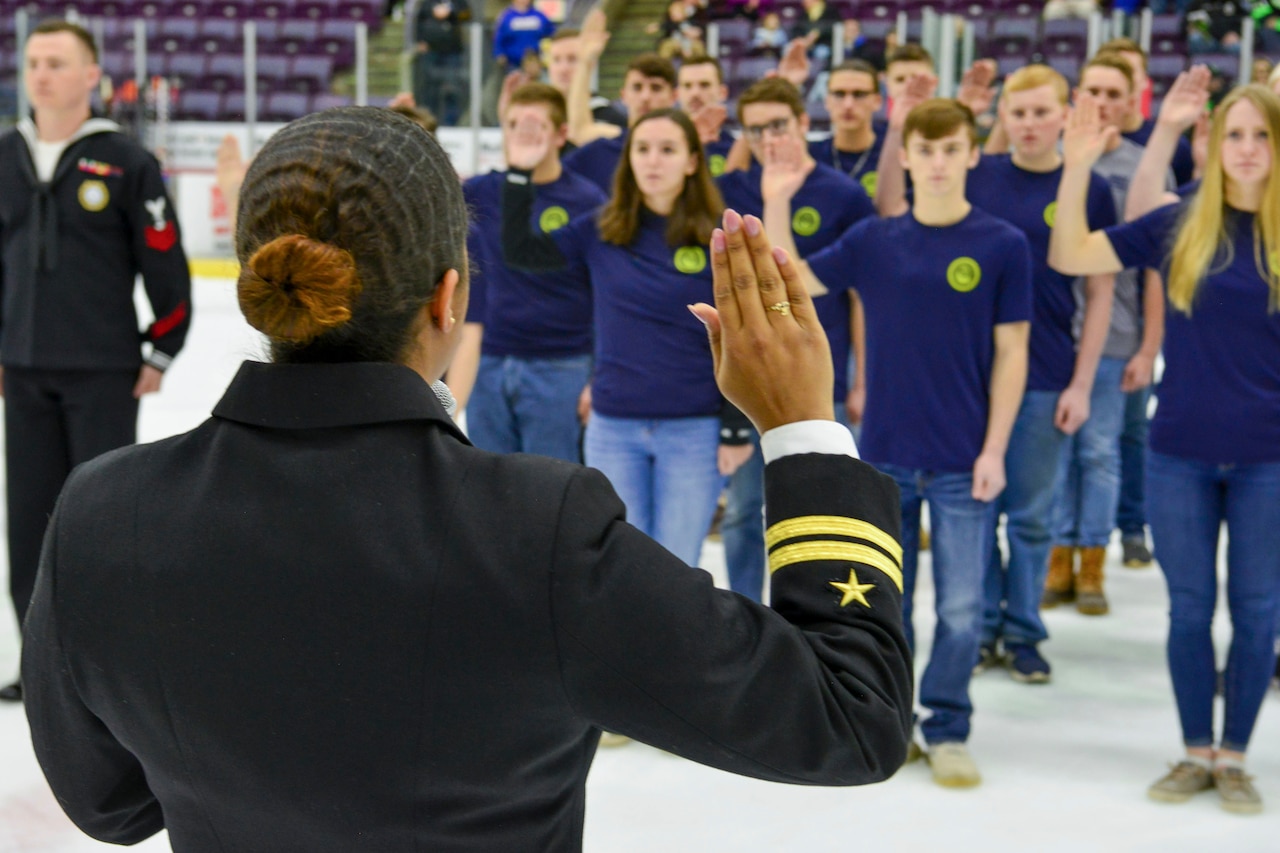 A sailor, shown from behind, raises her right arm and faces a group of young people doing the same thing at an ice rink.