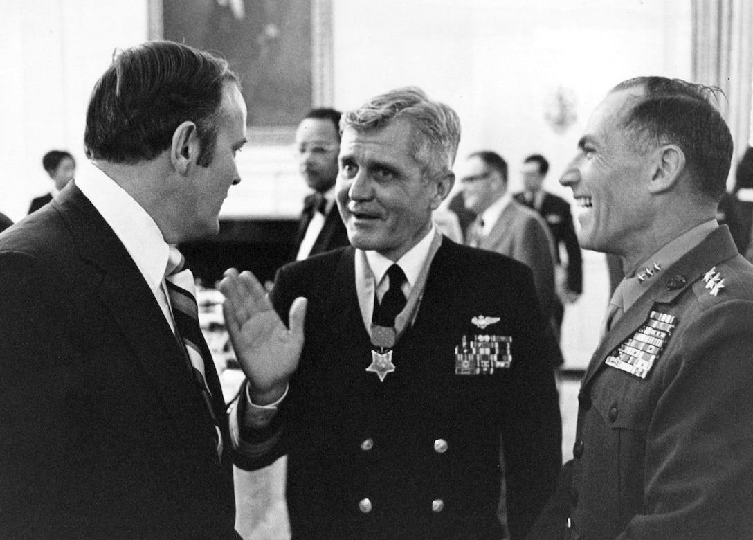 Three men, two wearing military uniforms, stand talking. The man in the middle wears a medal.