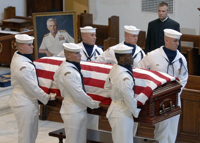 Six sailors carry a flag-draped casket. A portrait of the man who died is on display.