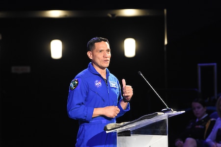 Hispanic man in blue jump suit stands in front of clear podium with microphone.