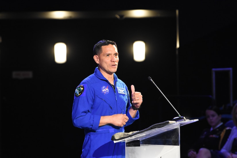Hispanic man in blue jump suit stands in front of clear podium with microphone.
