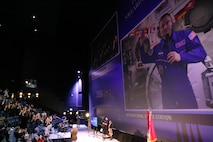 Many people in black tshirts stand with right hand raised looking at a blue screen with a man in a blue jump suit who is also raising his right hand.