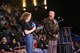 Girl with curly hair in black shirt and blue pants holds microphone standing next to man in green and kaki uniform at a black wire podium.