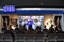 rows of people sitting in chairs watching a man in space on a large sized display.