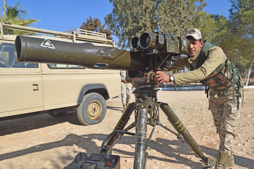 Jordanian Soldier stands with weapon on tripod, bent to look through the scope, with left hand on a knob
