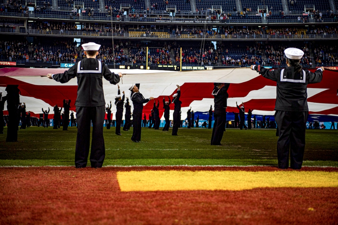 Sailors hold up a giant American flag on the field of a football stadium.