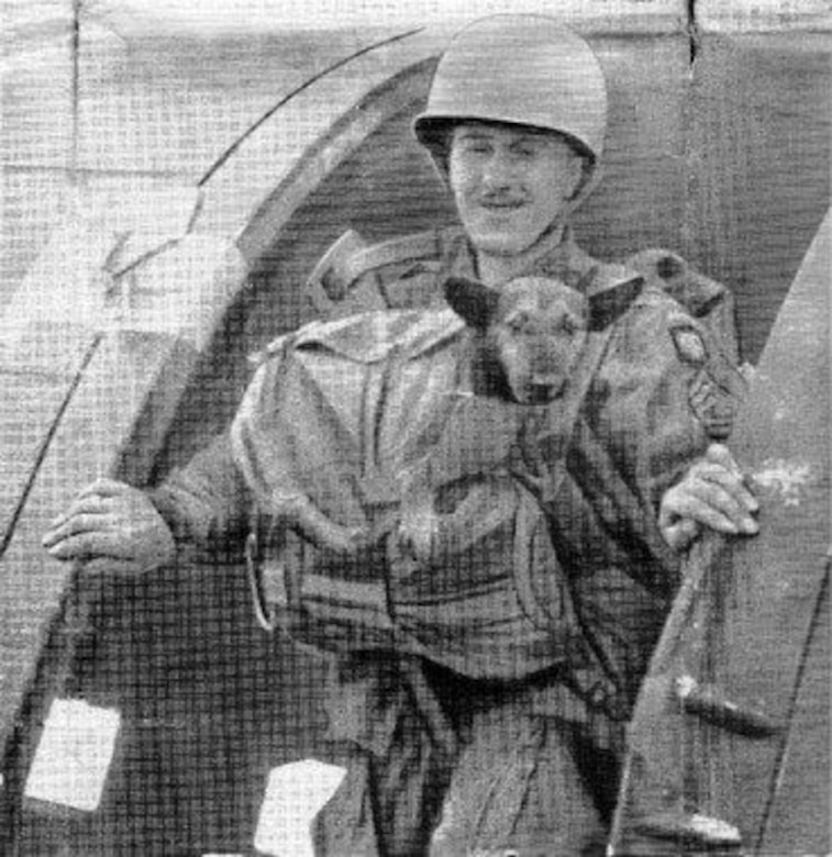A World War II paratrooper wearing a combat helmet and parachute gear has an 8-month-old puppy strapped to his chest. Both stand at the open door of an airplane.