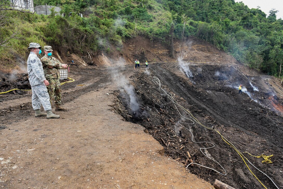 Two airmen wearing protective masks look at a smoldering patch of ground.