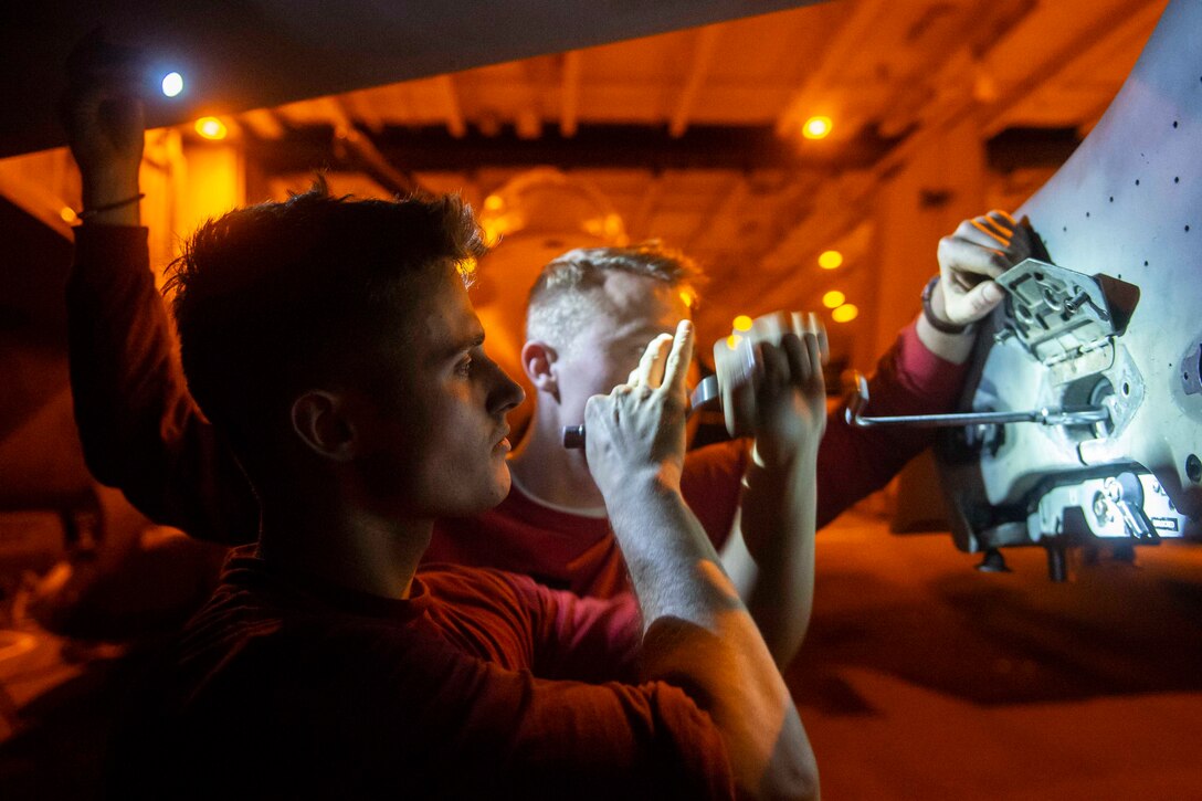 A sailor works on a military jet in the dimly lit hangar of an aircraft carrier.