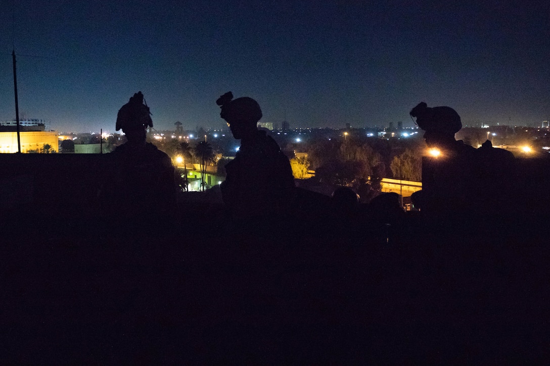 Three Marines wearing combat equipment are silhouetted against a city skyline at night.