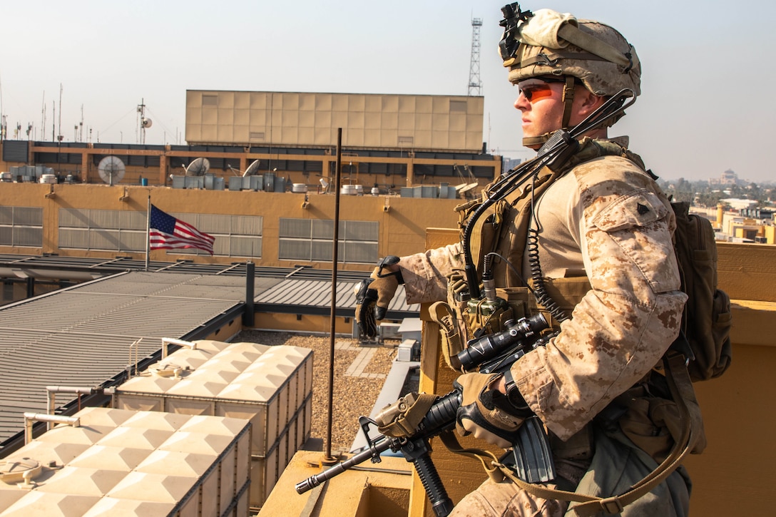 A Marine with combat gear stands watch on top of a building. A U.S. flag is in the background.