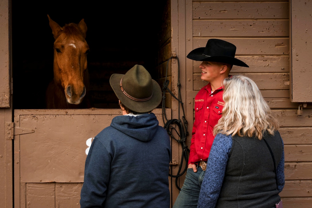 A Marine wearing a cowboy hat and two civilians look at a horse peering out from its enclosure in a stable.