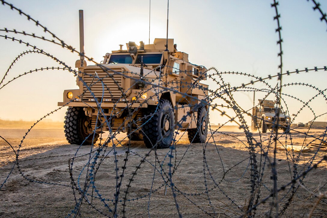 Two tactical vehicles drive on dirt ground, with looped barbed wire in front of them.