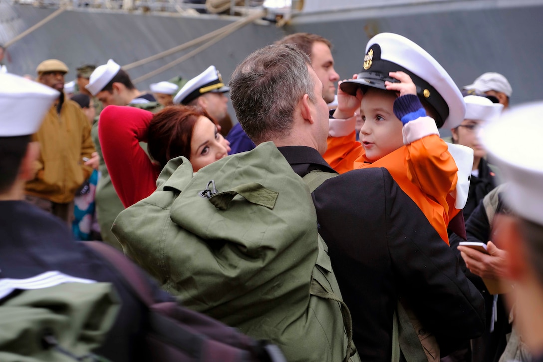 A child tries on a sailor's hat while being held in a man's arms.