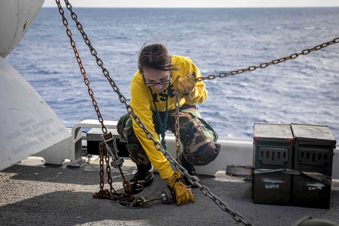 A kneeling sailor manipulates chains on a flight deck.
