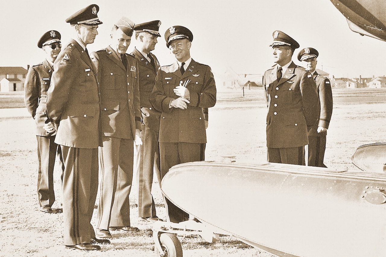 Several high-level military officials in dress uniforms and caps chat beside the wing of an airplane on an airfield.