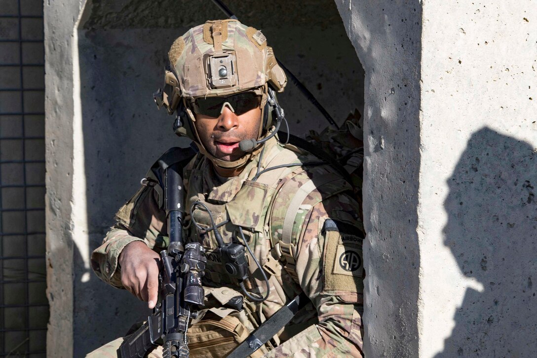 A soldier holding a weapon stands against a building exterior and looks out at his surroundings.